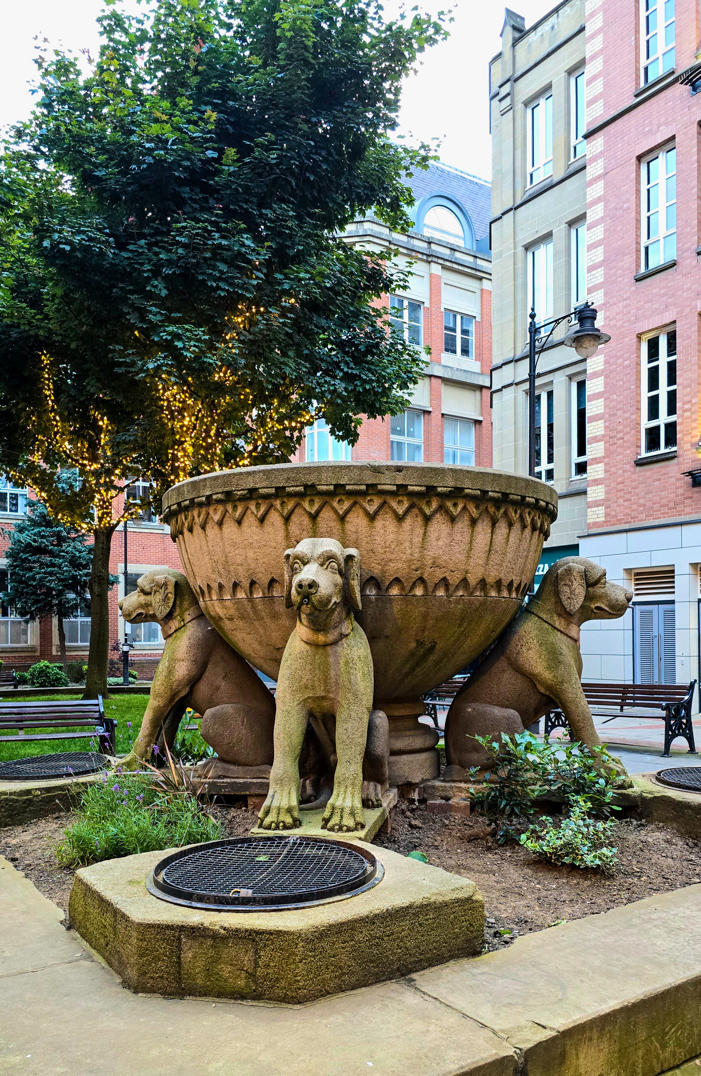 Dog sculptures supporting a large stone bowl fountain in a small city park in Leeds