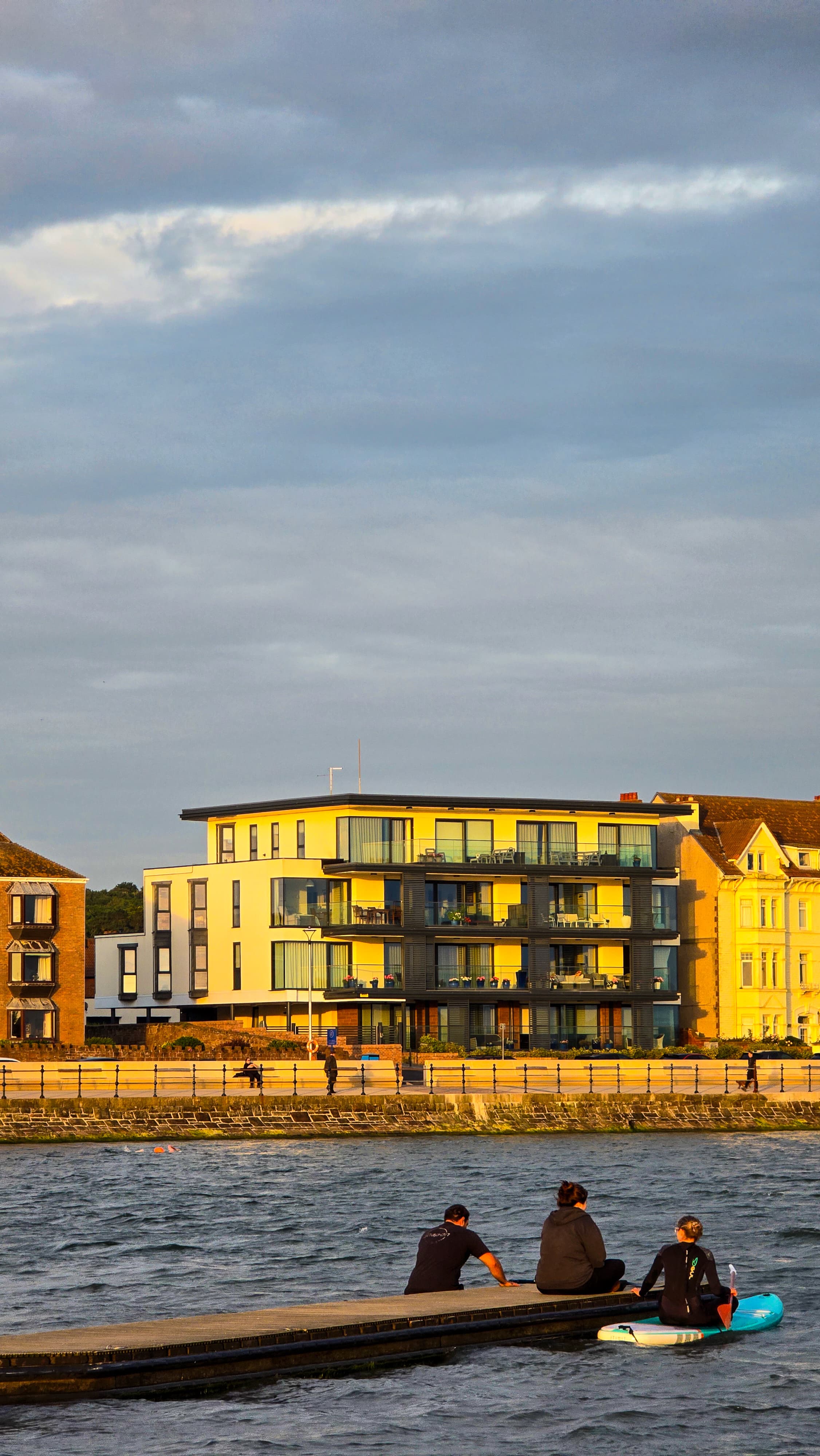 Three people resting on a dock at West Kirby Marine Lake with soft evening light on modern buildings across the water.