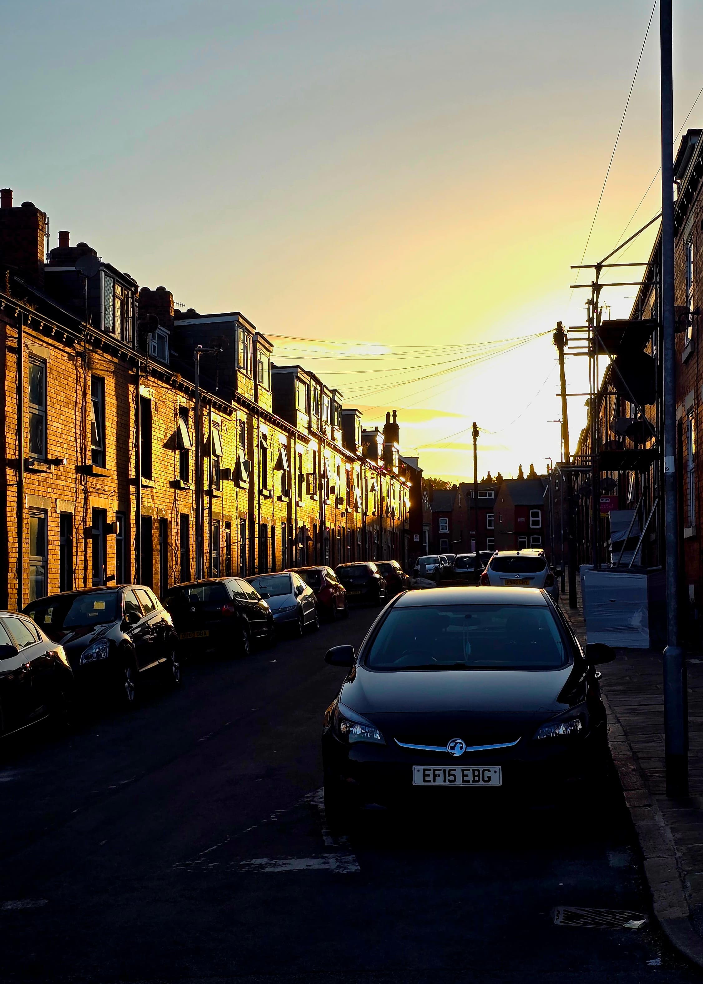 Golden sunlight falling on a quiet street in Leeds with brick row houses and parked cars.